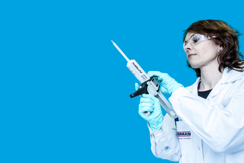 A female lab technician wearing a white lab coat, teal gloves, and safety goggles holds a dual-cartridge adhesive dispenser labeled “Araldite 2000” by Huntsman, against a solid blue background.