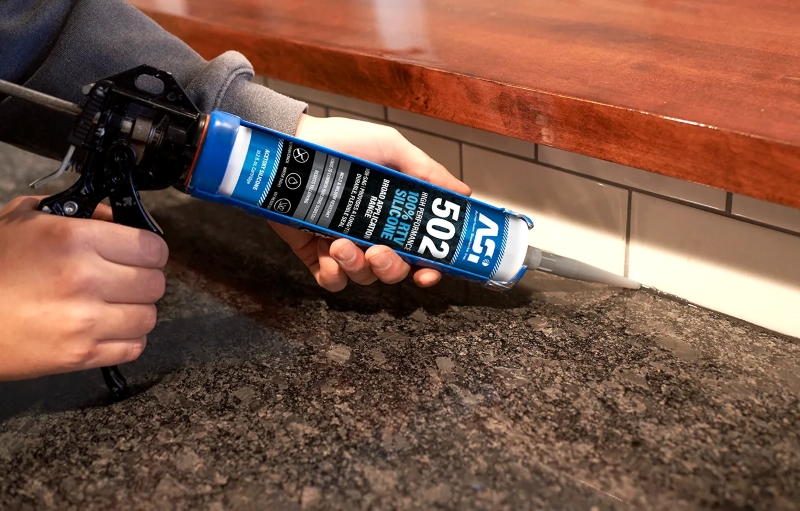 Caulking gun applying ASI 502 silicone along the joint between a dark stone countertop and a white tile backsplash