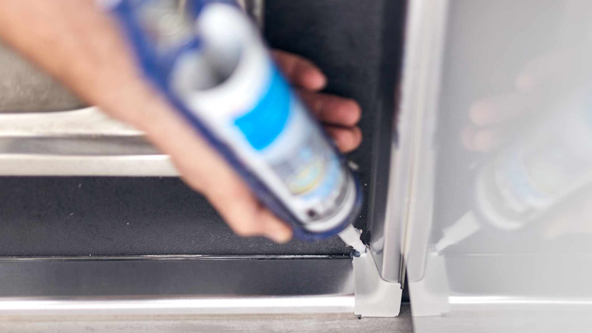 A hand holds a tube of structural adhesive, applying it to a seam between two metal surfaces.