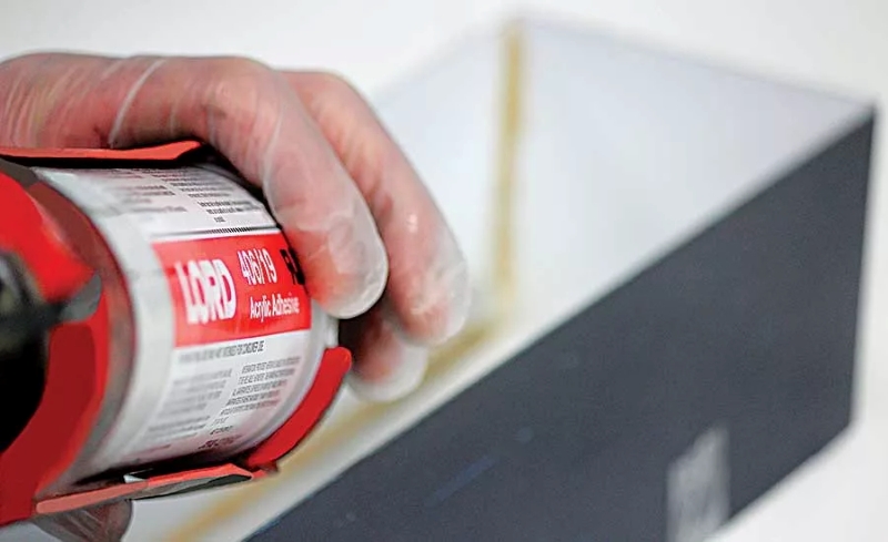 Gloved hand dispensing LORD 406/19 acrylic adhesive from a cartridge along the inside corner seam of a sheet metal box